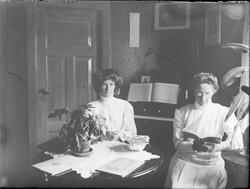 To kvinner sittende ved et bord i stue, en av kvinnene leser