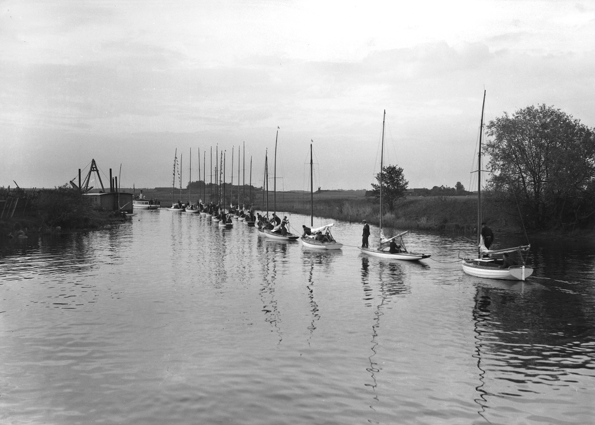 Ångslupen Tasse med motorlösa segellbåtar på släp genom Stångåns slingriga flöde mot sjön Roxen, där det sannolikt stundar kappsegling. Notera även stadens mudderverk till vänster. Dagen var den 8 juni 1935.