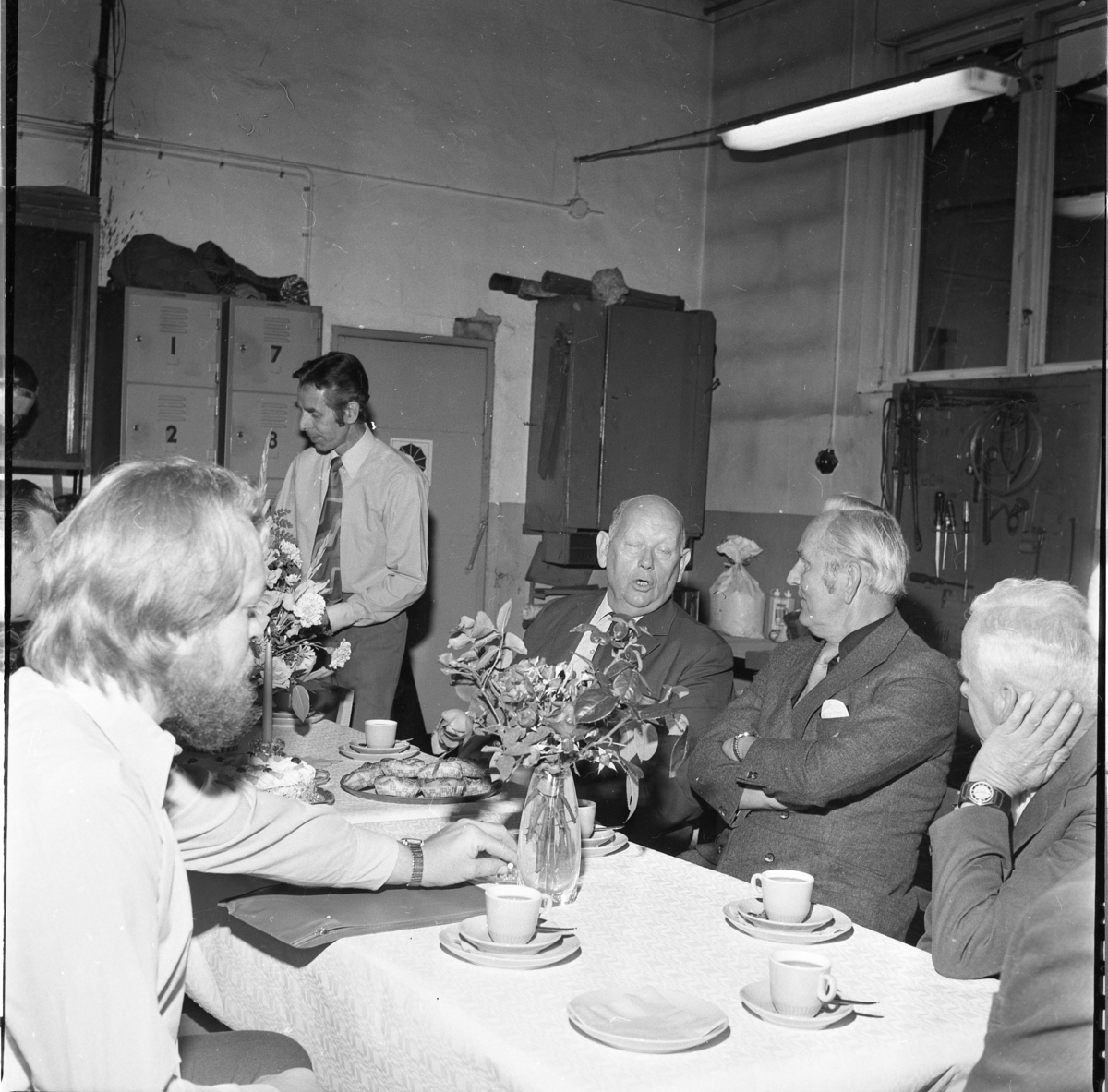 Män i samtal runt ett bord med kaffekoppar, bullar, tårta och blommor.