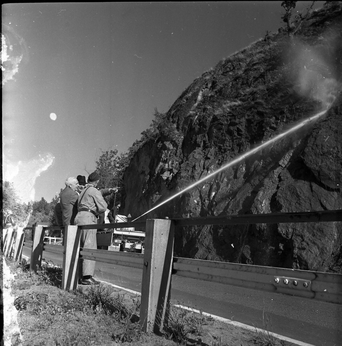 Rasberget vid länsgränsen 1961. En vattenkanon åker längst med en motorväg och har kanonen riktad mot en bergvägg. Vattnet träffar högt upp på berget och nedanför ligger rasmassor. Mot vägstaketet står tre herrar lutade, en av dem är polis.