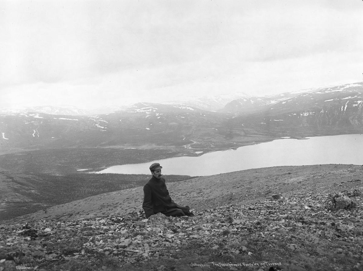 En mann har satt seg til på Trollsteinhøe med utsikt til Veodalen, Nedre Steinbuvatnet, Glittertind og Kvitingskjølen, 1916.