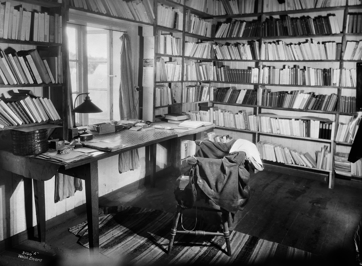 Del av Knut Hamsuns arbeidsværelse.  Klesplagg på stolen ved skrivebordet, veggene fulle av bøker, skrivebord plassert under vinduet.