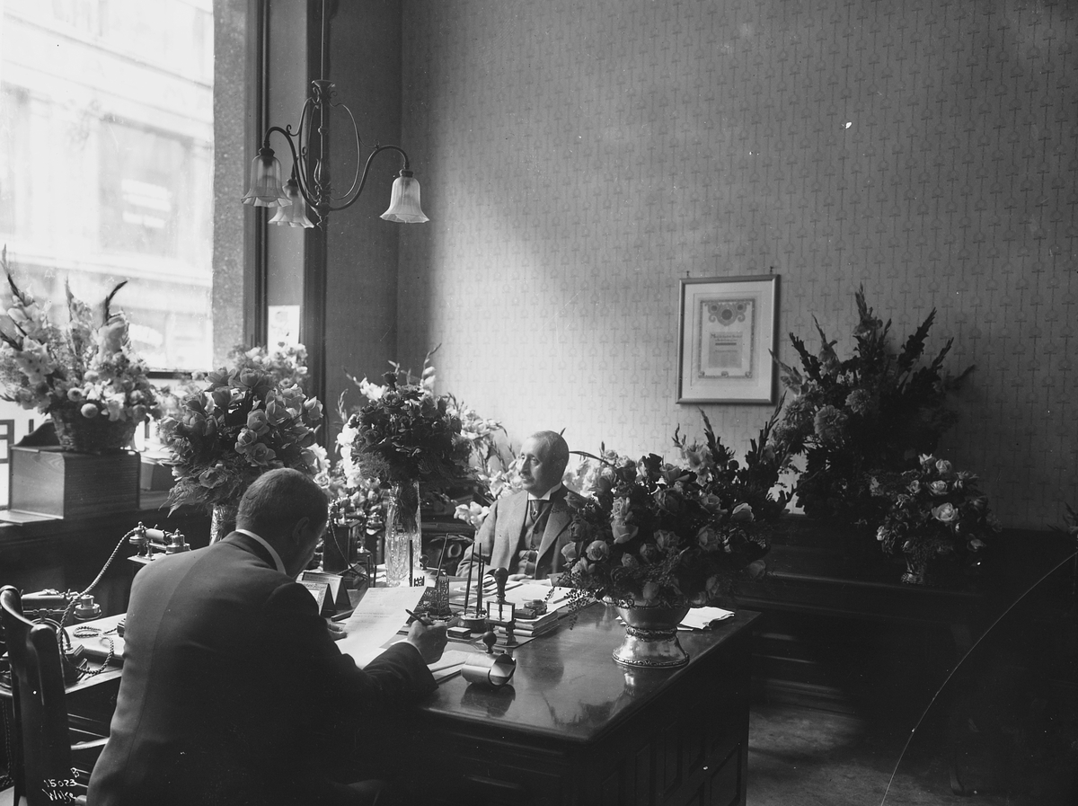 Kontoret bærer preg av festligheter og jubileum, de to herrene sitter omringet av blomsterdekorasjoner.