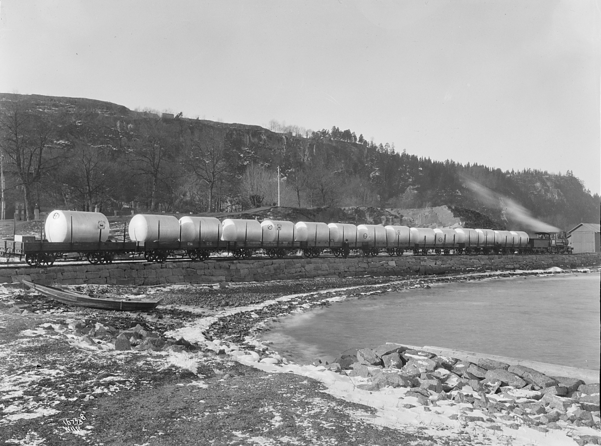 Godstog kjøre med mange vogner lastet med ant. aluminium. Norsk Aluminiumindustri. Robåt ligger på land langs toglinjen. Fotografter 1923.
