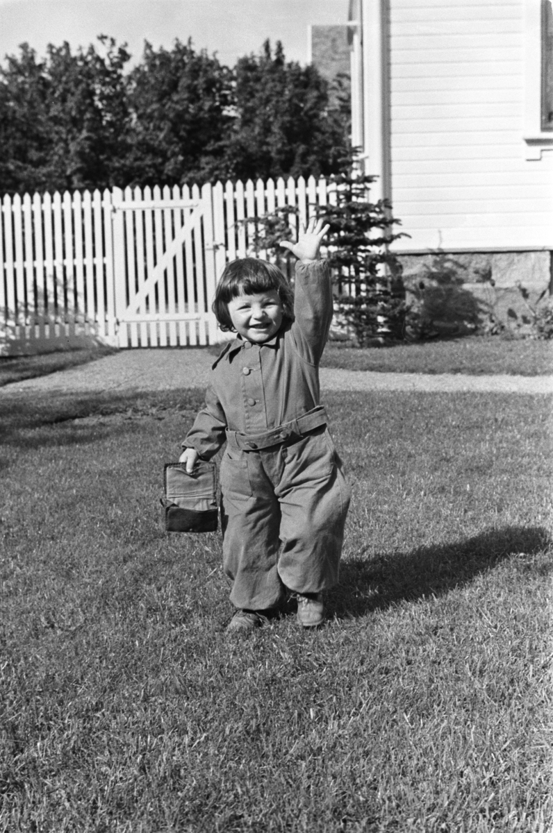 Blid liten jente i hagen, bærer på en veske.  Del av huset og gjerdet i bakgrunnen.