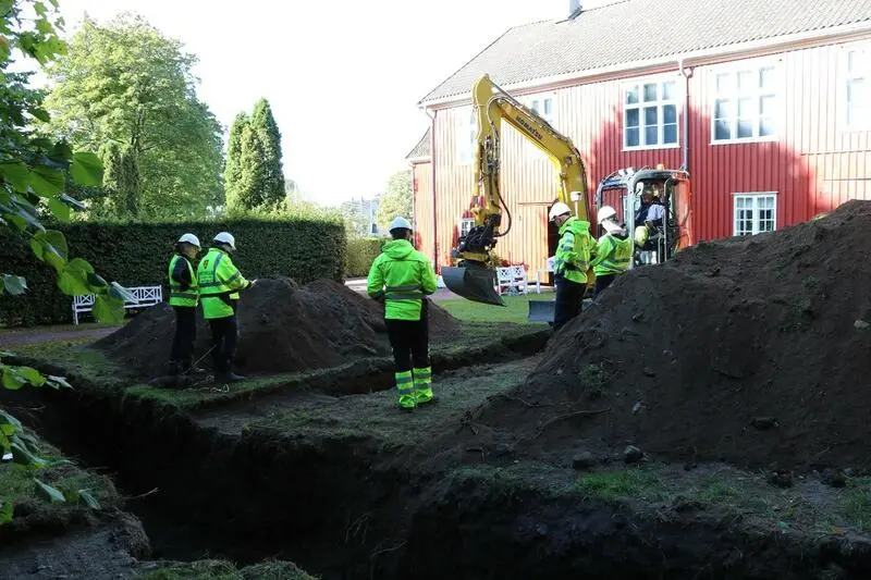 Graving i bakken. Vi ser en gravemaskin i høyre del av bilder, og flere mennesker med gule refleksvester og hjelm rundt - og i hullet.