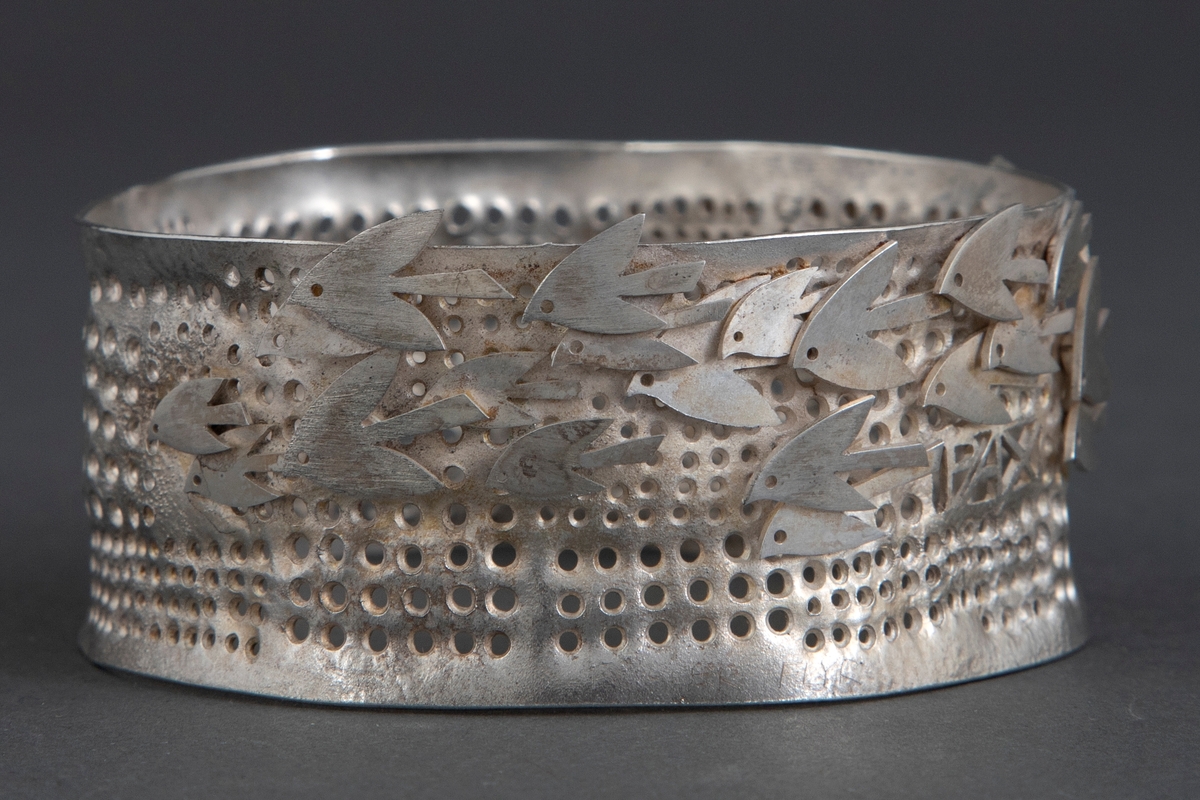 Tilnærmet sirkulær armring i sølv. Overflaten er perforert, og dekorert stiliserte fugler.