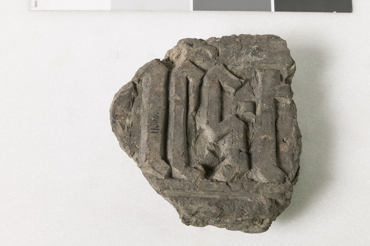 Et lidet Fragment af en middelaldersk Ligsten, med Minuskelindskrift, hvoraf blot et Par Bogstaver kunne læses. Stl: 13,7 cm, Stb: 13 cm, Sth: 6,4 cm. Vekt: 912 g. 