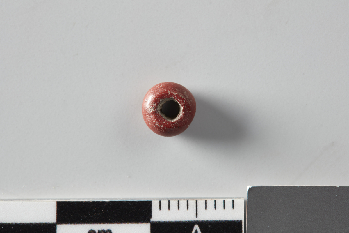 Mørk rød glassperle funnet i S318.  Ett av hullene i perlen er noe større enn det andre.