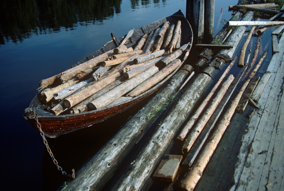 «Fetsund-båt», fylt med bruddstykker av tømmerstokker, muligens etter «reingjøring» av retteanlegget ved lensene (jfr. SJF-F.014540 og SJF-F.014541) Dette var i hvert fall virke som det ikke hadde noe for seg å forsøke å «soppe» (bunte) for buksering over innsjøen Øyeren. Fetsund-båten fantes i to utgaver – en klinkbygd, der bordgangene var lagt med et visst overlegg hvor det var innsatt klinknagler, og en der bordgangene var spikret på spantene slik at den ytre huden ble noenlunde glatt. Det siste kunne være en fordel når farkosten skulle gå i et lenseanlegg der det flytende tømmeret kunne bli en påkjenning på de overlappende skjøtene mellom bordgangene. Dette later til å ha vært en klinkbygd modell. Båten er tjærebredd, slik de fleste robåtene i lensemiljøet var. Bare formannsbåtene var gråmalte. Lenge var det særlig Peder Olsen Strandbakken (1827-1912) og sønnen Oskar Pedersen Strandbakken (1888-1965) som leverte robåter til lenseanlegget. Andre leverandører var Anton Martinsen Linnerud (1867-1947), Ole Larsen Strandbakken (f. 1862), Karl Lilleberg (1884-1973), Ole Stubberud (1887-1981) og Dinius Stenerud (1894-1983). Ut fra et fotografi som dette er det naturligvis vanskelig å ta stilling til hvem som kan ha vært mester for akkurat denne båten.