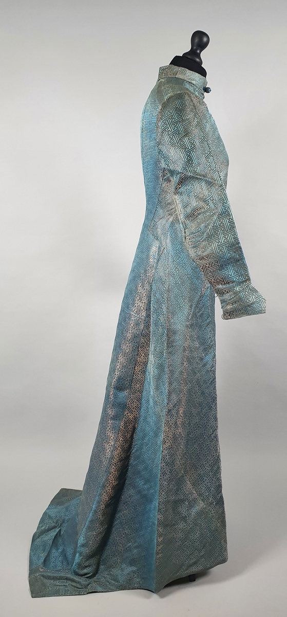 Turkis silkekjole med lange ermer og slep. Knapper diagonalt over brystet på venstre side. Föret med turkis silke.