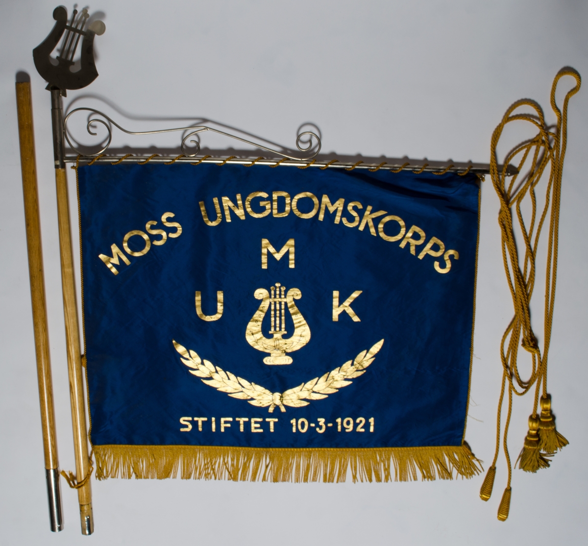 Blått banner med galleri, toppspyd, bærestang, bardunsnor, styresnor. Banneret er kantet nederst med gule frynser. Midtstilt motiv med stemmegaffel og krans under.