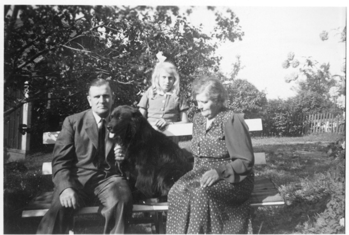 En man och en kvinna sitter på en trädgårdssoffa med en hund mellan sig, en liten flicka - dottern - står bakom dem.