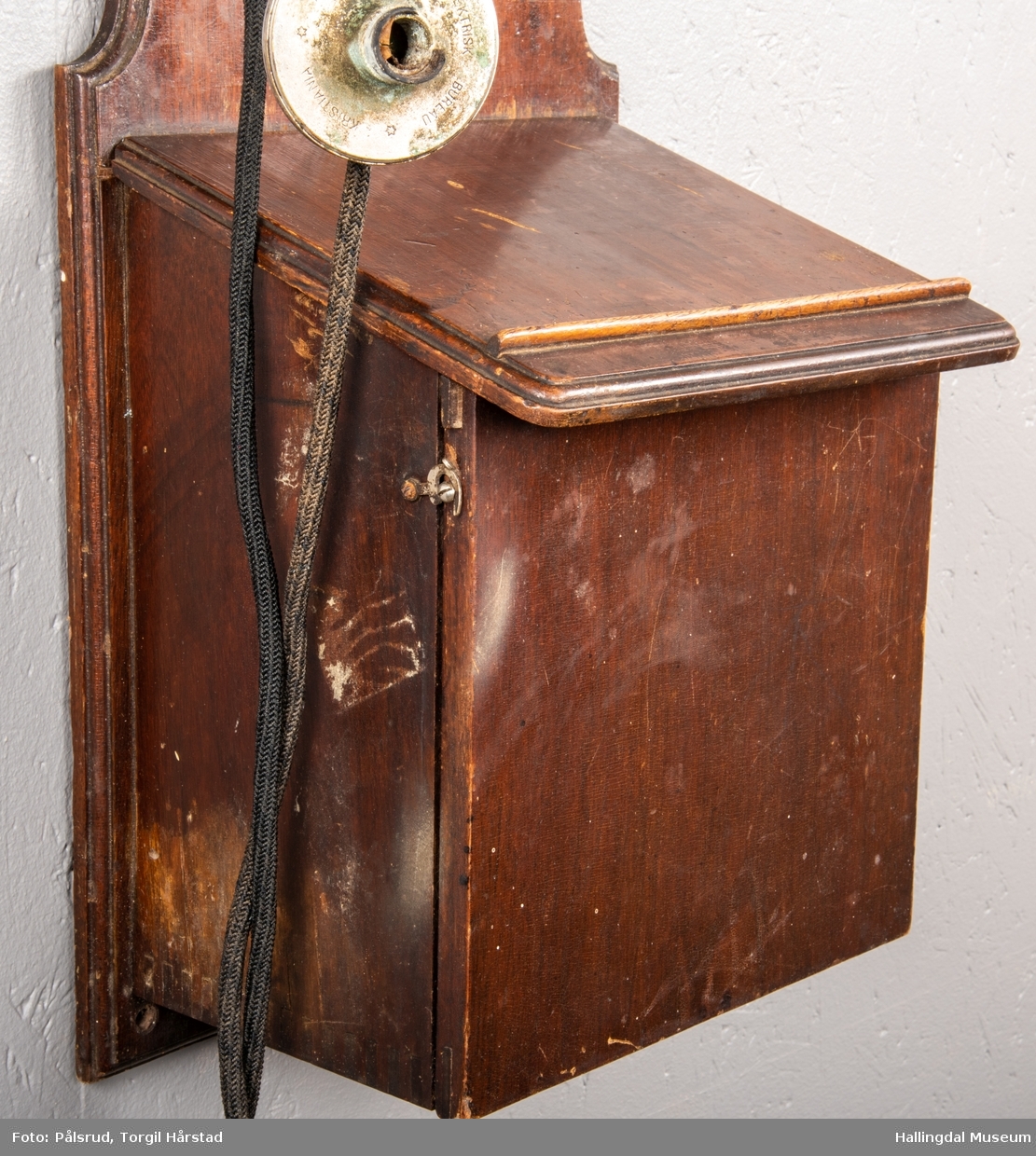 En veggtelefon i tre med to ringeklokker i blank metall øverst på boks. Svart leding koblet til telefonrør og sveiv med svart plasthåndtak på motsatt side. Flere ledninger på baksiden. Nederst en boks i tre for oppbevaring av to batterier produsert av Norsk Akkumulator Co. A/S - påskrevet dato 1953. Telefonen har tilhørende skriv med regler for telefonbruk for Tunhøvd - Næstelefon, med dato 1918. 