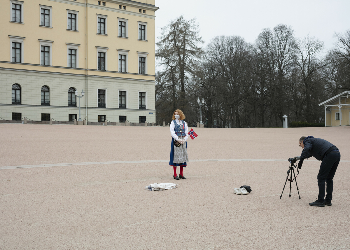 Kvinne iført bunad og maske, står på Slottsplassen med et norskt flagg sammen med en fotograf.