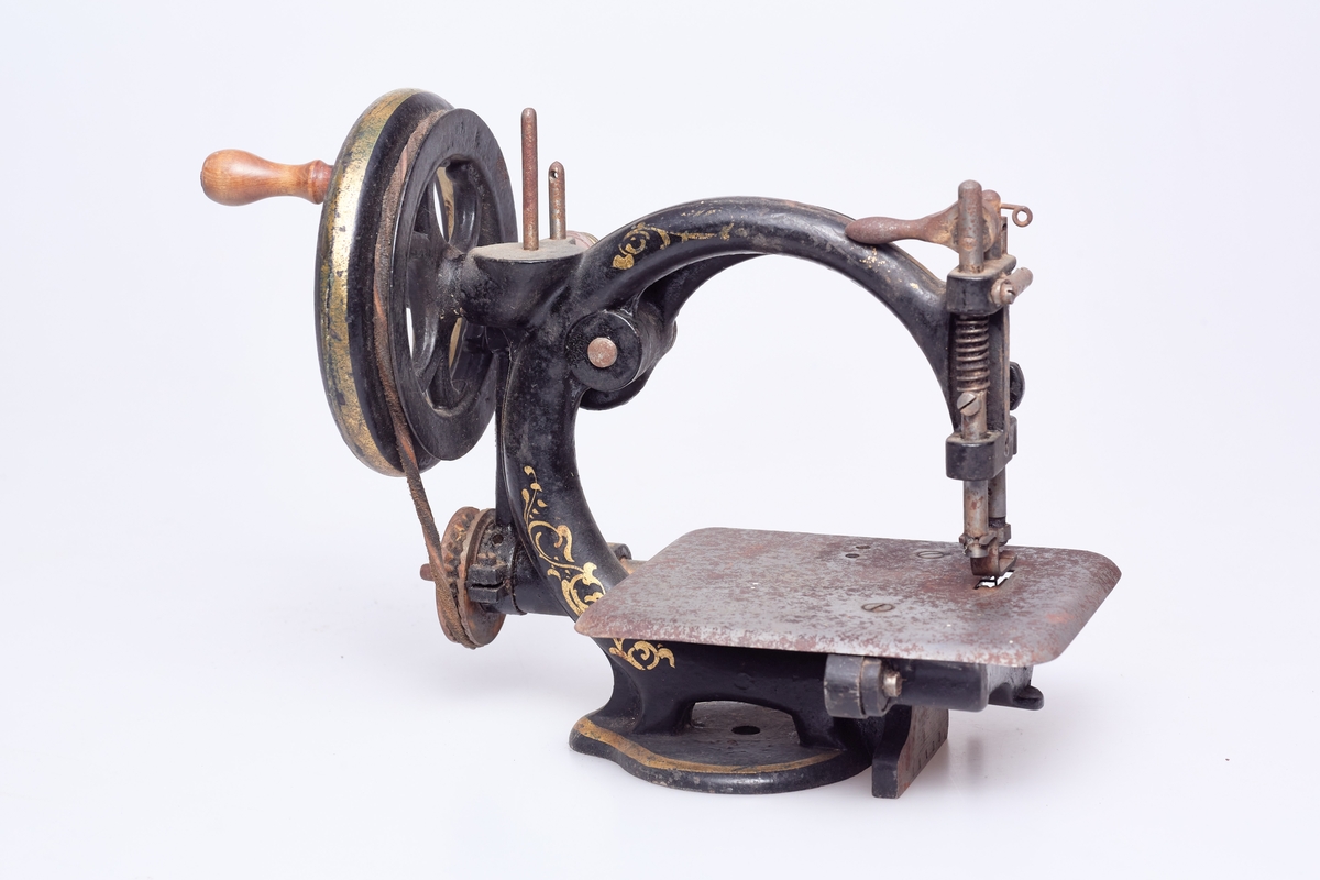Symaskin for håndkraft. Symaskinen er enkelttrådet. Rødmalt kasse for symaskinen av gran med skyvelokk. I kassen ligger mange reservedeler og 3 nåler.  Noe er innpakket i avispapir fra 1899.