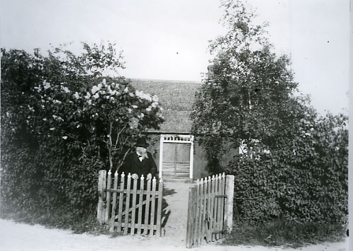 Författaren, läkaren och folklivsskildraren August Bondeson vid grinden framför Fågelboet, hans föräldrahem i Vessigebro. Tomten hägnas av syrénhäckar och en ung björk står vid grinden.
