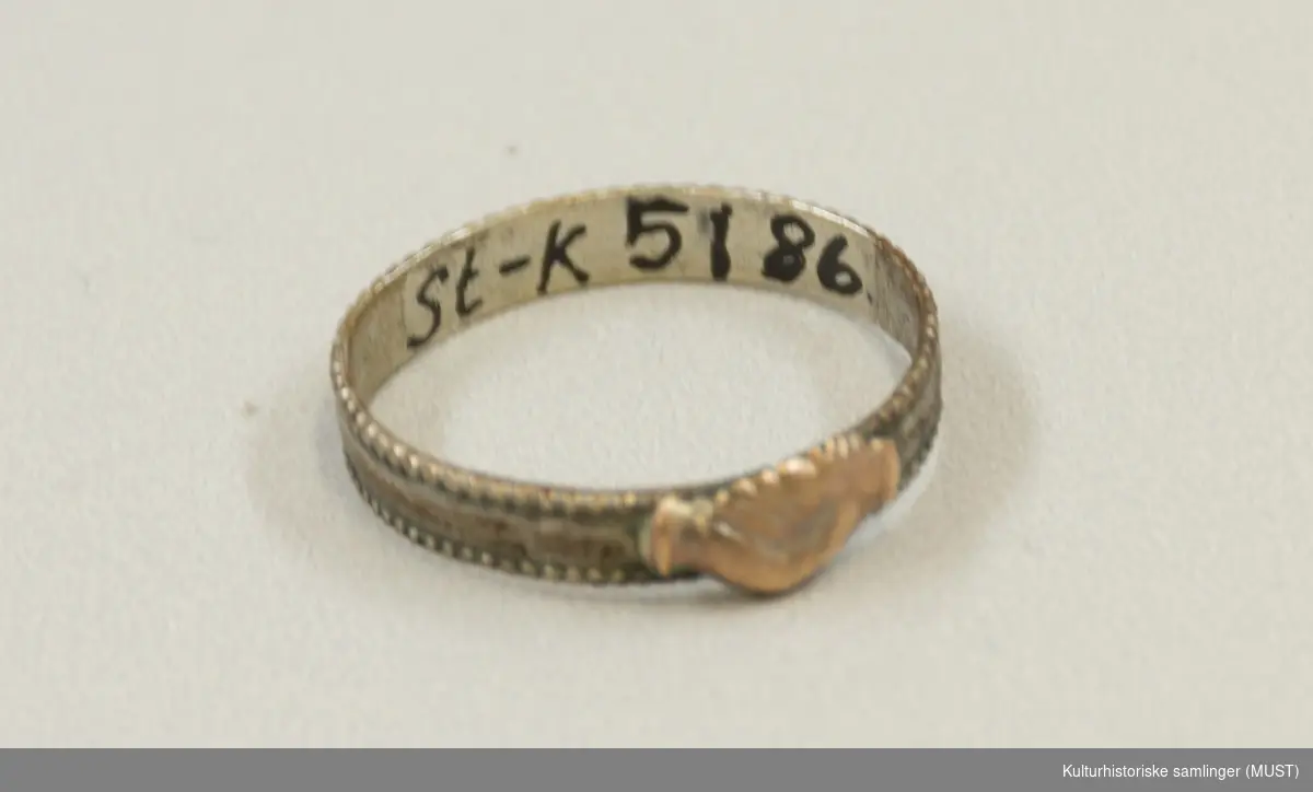 Glatt, flat ring med små, graverte border langs kantene. Påloddet ornament i
kobber bestående av to hender som holder hverandre.