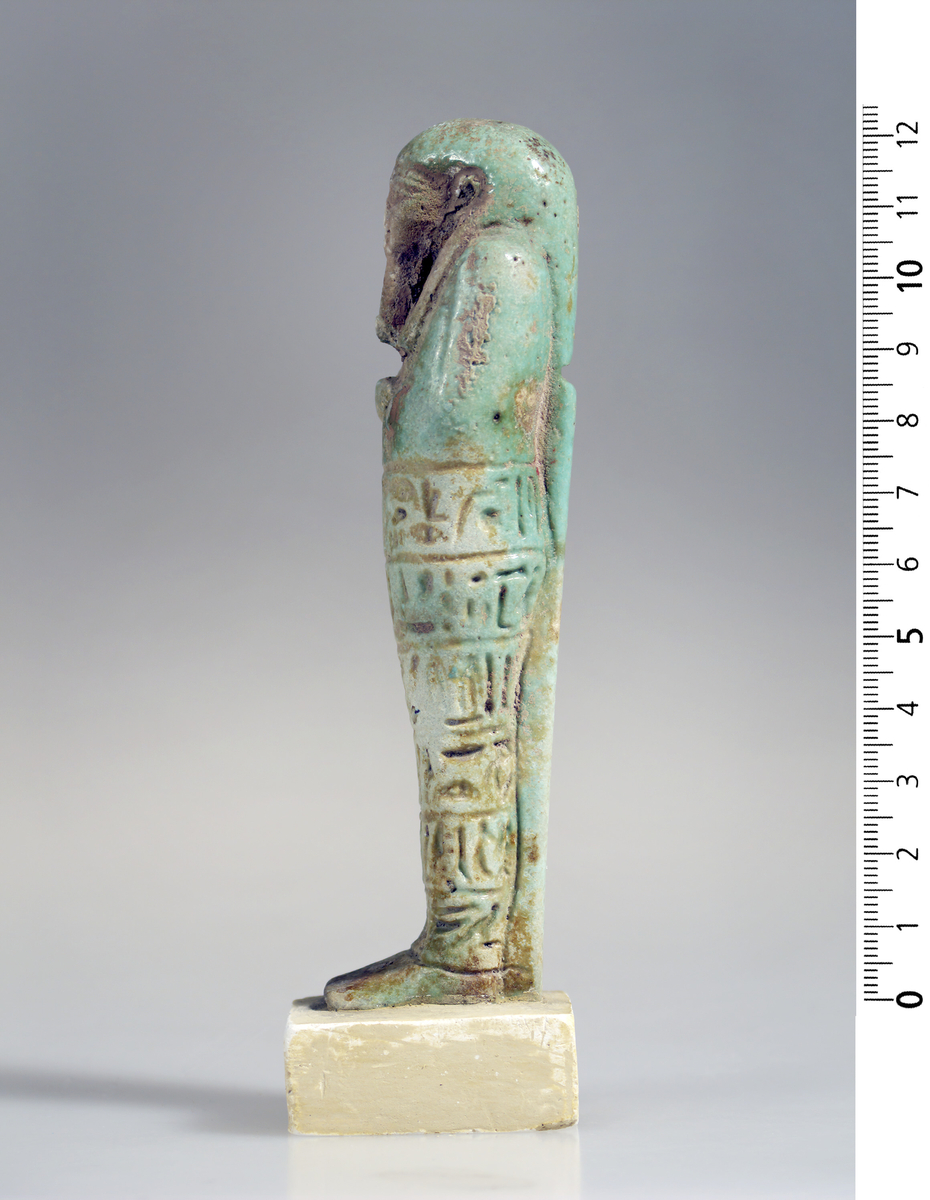 Ushabti i grønn fajanse med hieroglyfer foran. 12,5 cm høy.
Fajanse