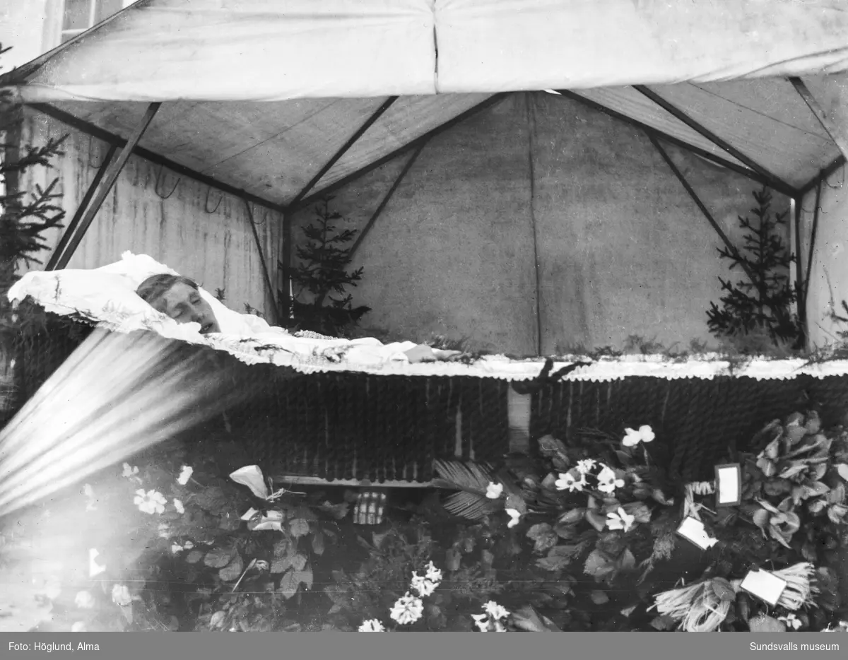 En ung kvinna i öppen kista med blommor framför utomhus. Bakom står något slags tält.
