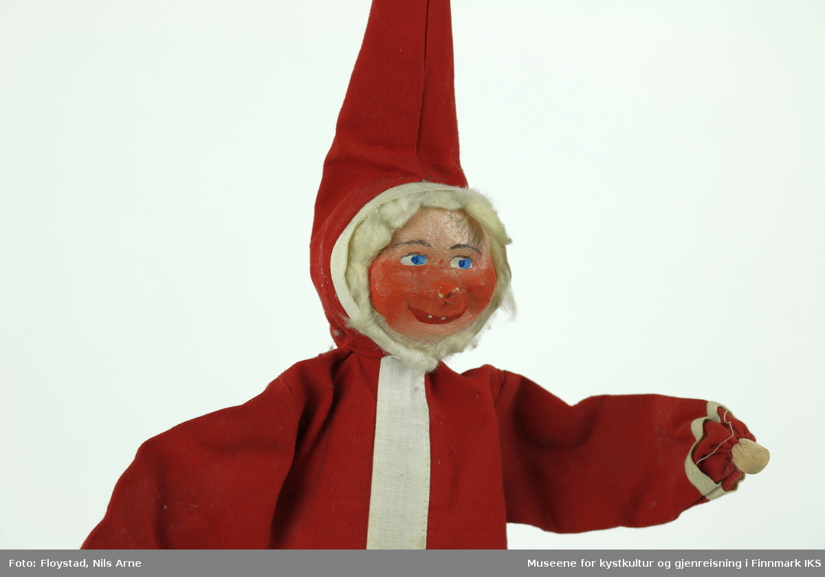 En julenisse laget av papp og tøystoff. Tøystoffet er i fargene rød og hvit, bortsett fra hendene til nissen der har tøystoffet en hudlignende farge. Ansiktet til nissen er laget av pappmasje også er det påmalt øyenbryn, blå øyner og munn. Rundt hodet er det pålimt bomull. 