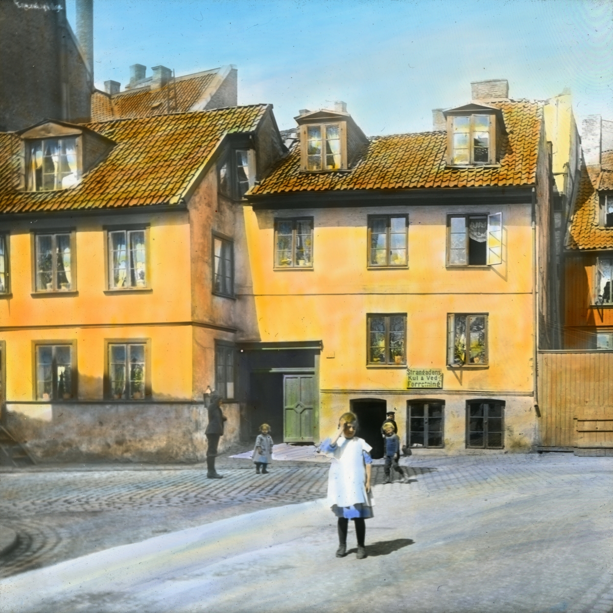Håndkolorert dias. En jente kledd i kjole står på gata foran en bygård i Vika, Oslo, i 1904. I bakgrunnen står tre andre små barn og en ung gutt. På bygården henger et skilt for "Strandgadens Kul og Ved-Forretning".   