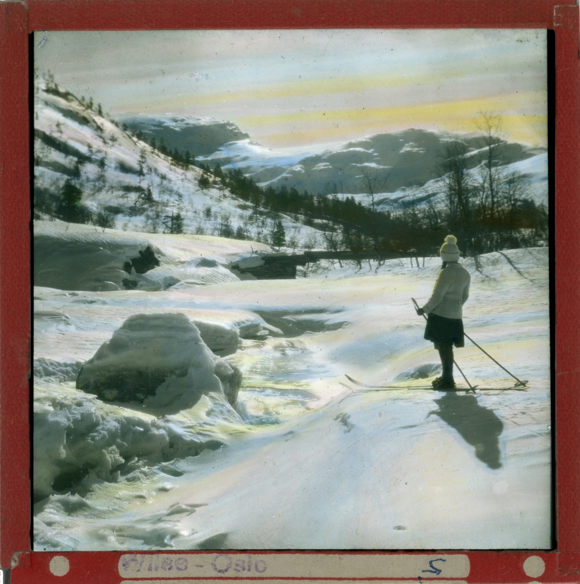 Håndkolorert dias. En kvinne på ski nyter utsikten over et vinterlandskap med fjell og skog.