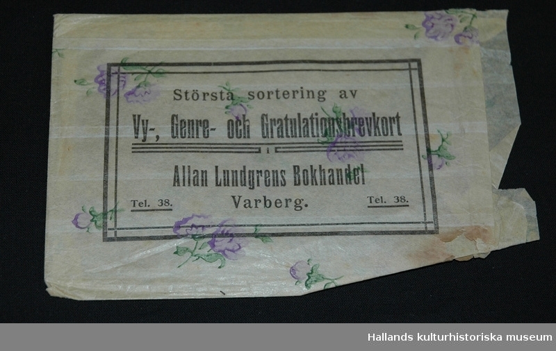 Påse i papper. Vit bakgrund med lila blommor. Märkning: "Största sortering av Vy-, Genre- och Gratulationsbrevkort, Allan Lundgrens Bokhandel, Varberg, Tel. 38". 