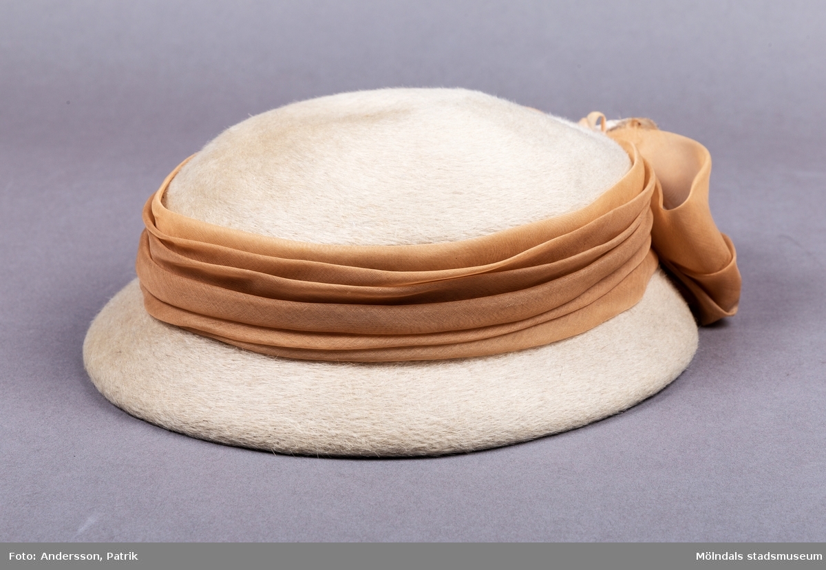 En damhatt i kamelhårsliknande mjukt material. Hatten är ljusbeige i färgen. Den är platt på ovansidan och har ett medelstort brätte. Runt hattens kulle finns ett beigefärgat sidenband som avslutas i en stor rosett på hattens baksida. På rosetten finns en rosa blomma i siden och sammet tyg. 
På insidan har hatten ett ribbsband med en fastsydd etikett. På etiketten finns tillverkarens namn Ateljévåningen Hattbazaren. Den finns ett gummiband fastsytt i hattens ribbsband, för att hålla hatten på plats vid användning.