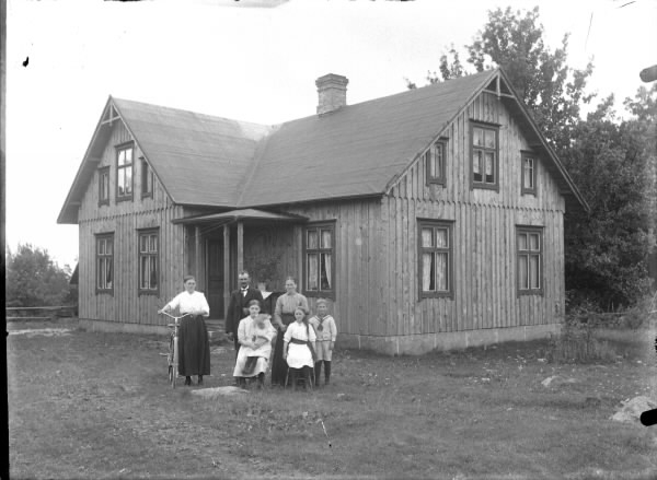 Familj med fyra barn och en kvinna med cykel framför ett omålat bostadshus med papptak och skärmtak över ingången i husvinkeln.