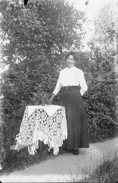 Kvinna står på en grusgång i trädgården vid ett bord med virkad duk.