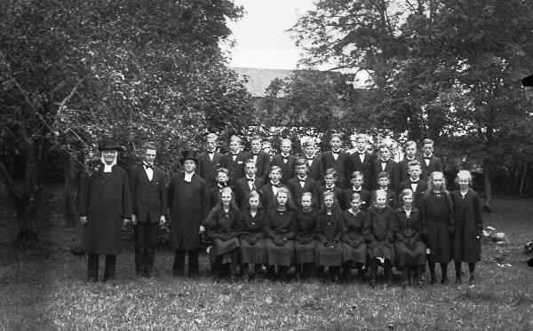 Mörkklädd konfirmandgrupp i en trädgård med präster i höga hattar till vänster, med en yngre man emellan sig.