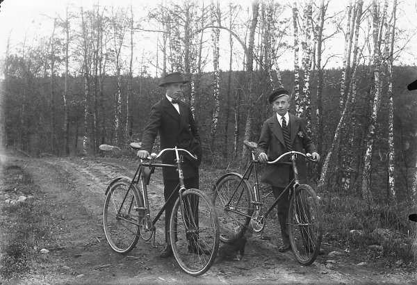 Bröderna Manne och Torsten Möller står i finkläder med sina cyklar på en grusväg kantad av unga björkar. Cykelstyrena är bockade nedåt.
