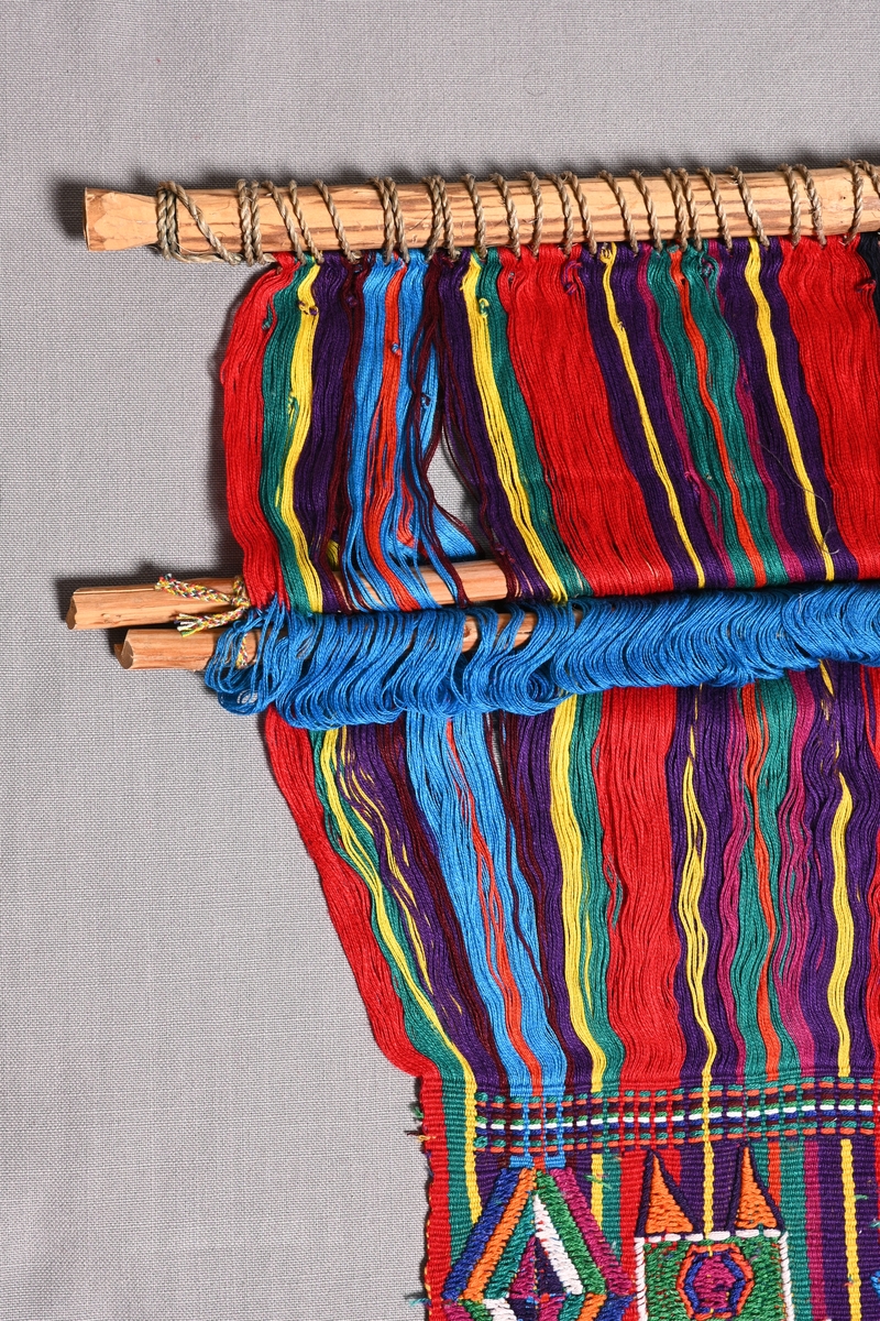 Vävuppsättning för ryggbandsvävning med halvsolv och skälblad (skälstock). Från Nebaj, Guatemala.
2-trådigt bomullsgarn i varp och otvinnat bomullsgarn i inslag i randig varprips, med inplockat mönsterinslag i flera olika färger.