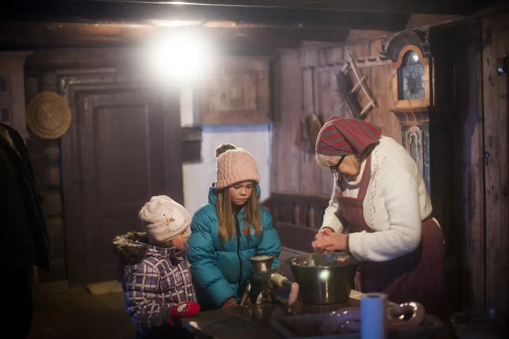 Bildet viser et eldre dame som stapper pølser. Det står to barn og ser på.