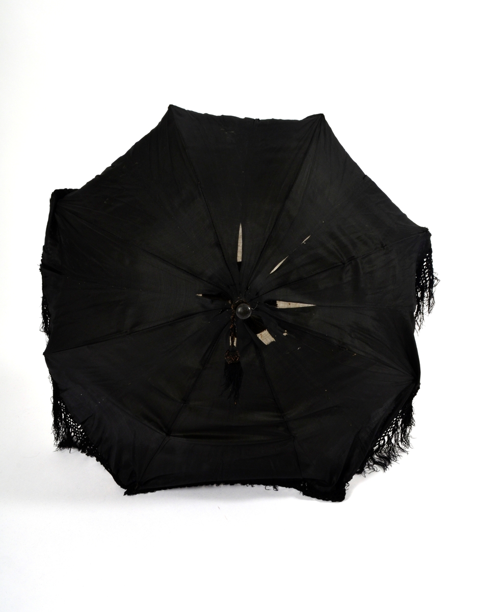 Parasoll i svart silke med frynserbord langs nedrekanten. Fóra med kvitt silketøy. 8 spiler. Stanga er utskåre i svart tre.