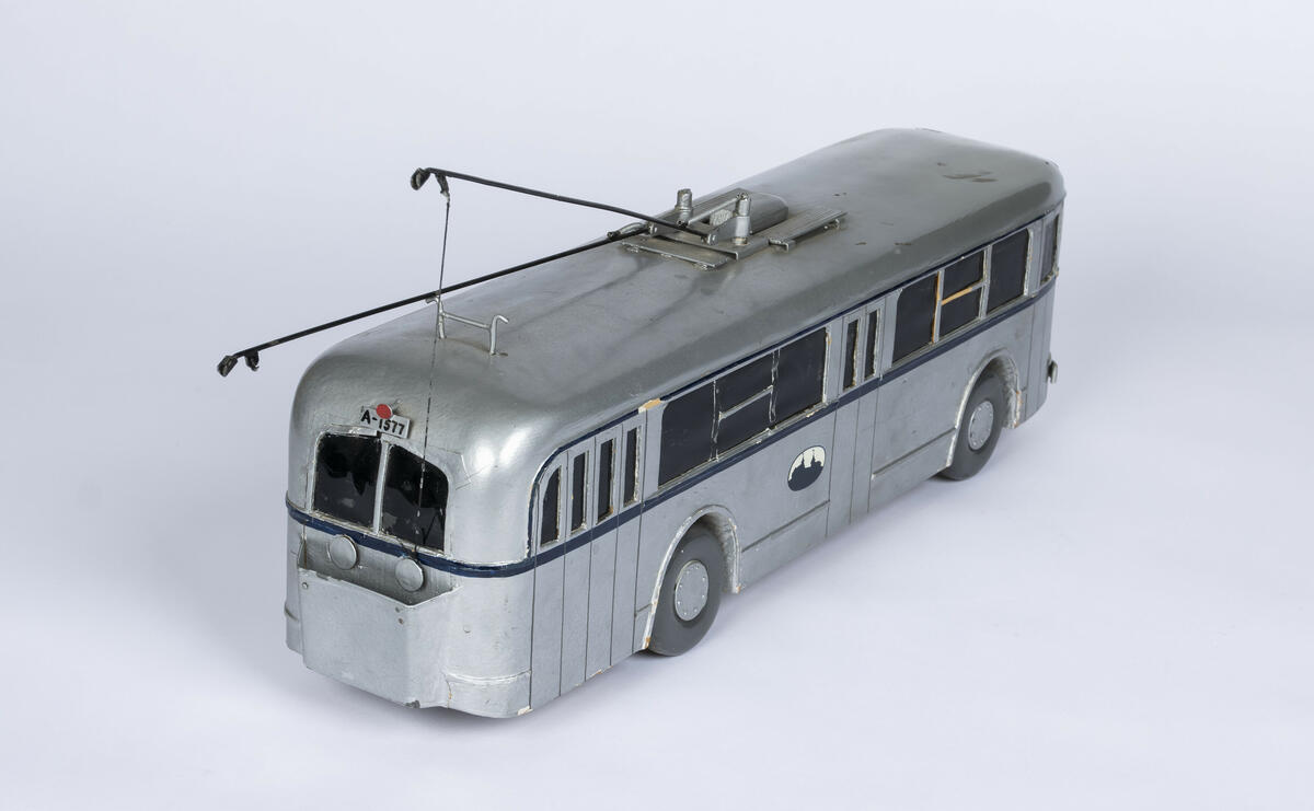 Modell av tre av buss A-1577. Bussen er farget i en sølvfarge med en mørk blå stripe på midten som går rundt hele modellen. Vinduene er farget sorte og de fire dekkene av tre er i en mørk grå farge. Bussen har tre inngangs- og utgangsdører. Denne modellen har to trolleystenger med kontakssko på enden og to trolleystenger som er festet til to snorspoler på baken av bussen. På enden av taket er det også plassert kroker for å feste trolleystengene når når ikker er i bruk.