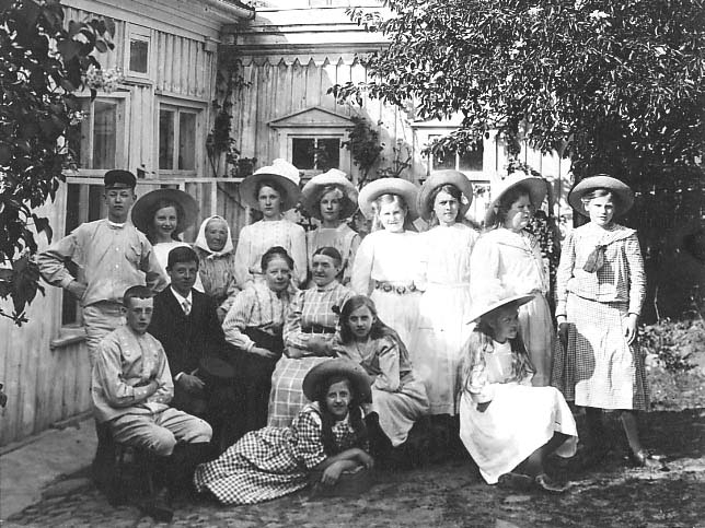 Elever, lärare och hushållerska vid fröken Valborg Francks privatskola i Gränna. Det står och sitter i trädgården utanför skolan.
