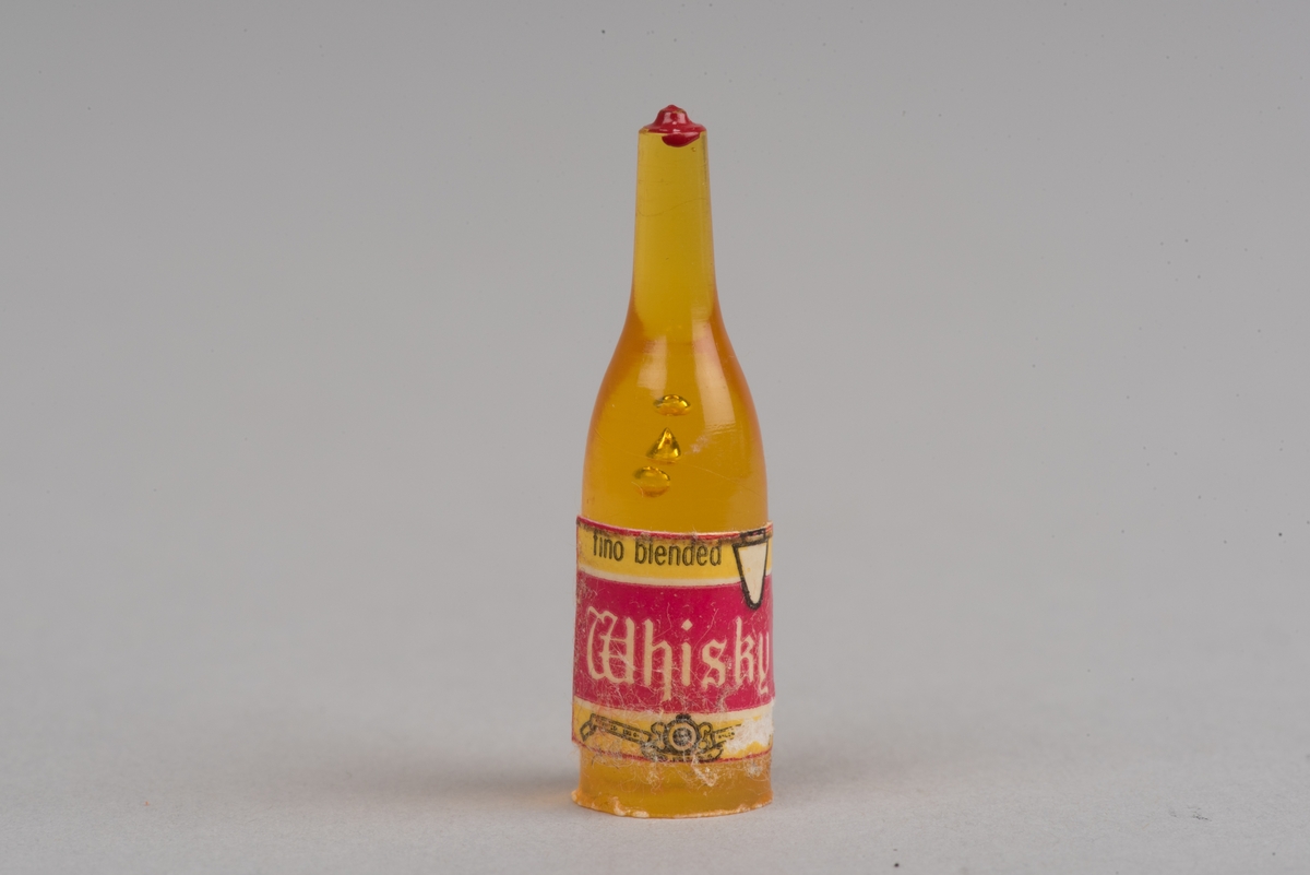 Dockskåpsinredning i form av en whiskyflaska i plast med pappersetikett.
Flaskan är gul med en röd kork. På etiketten framgår att det ska vara en whiskyflaska.