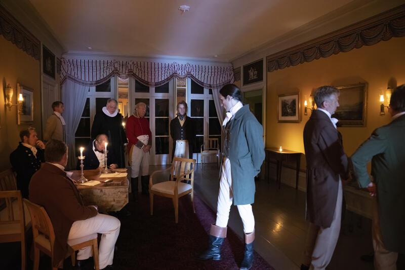 Foto fra hagestuen i Eidsvollsbygningen, fra filminnspilling. En samling av menn i kostymer gjenskaper stormannsmøtet 16. februar 1814
