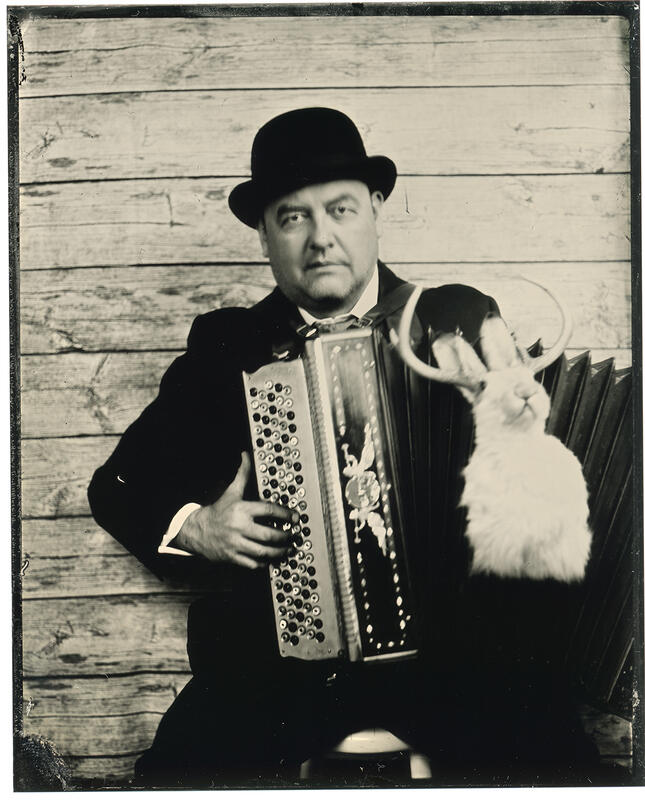 Fotografi av musiker Carsten Stiansen med hatt og dress, sittende på en krakk med trekkspill på kneet mot en vegg og en utstoppet, hvit kanin med horn i forgrunnen