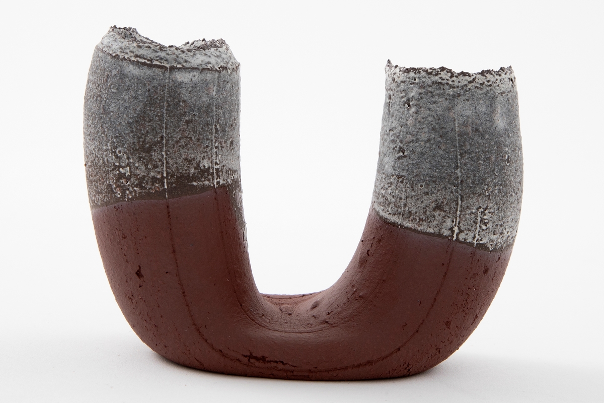 Vase i matt rødbrun terrakotta. U-form med riller og åpne ender med ruglete render. Hvit halvtransparent glasur på begge ender og på innsiden av munningene.