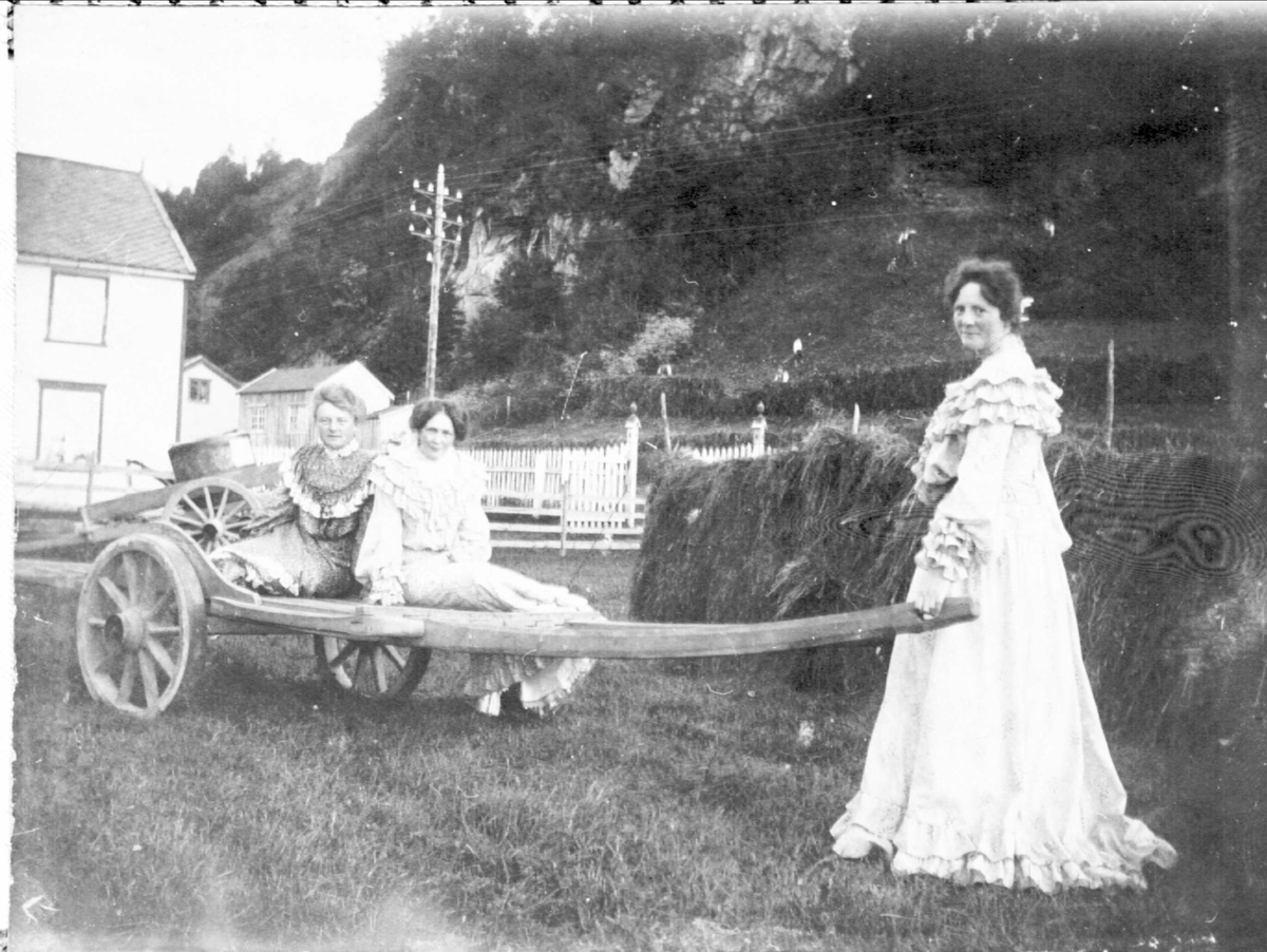 Tre kvinner i fine kjoler med hestekjerre, fotografert foran en hesje.