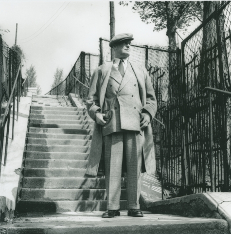 Maurice Chevalier i tweeddress og frakk fotografert i en trapp. 