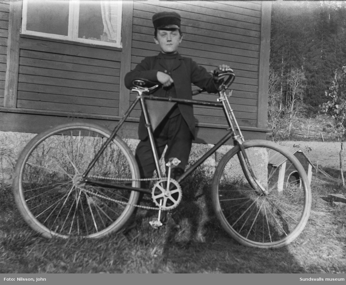 En pojke i skärmmössa står med en cykel vid husknuten till Roken 1:3 i Laggarberg.. Hundkoja i bakgrunden.
