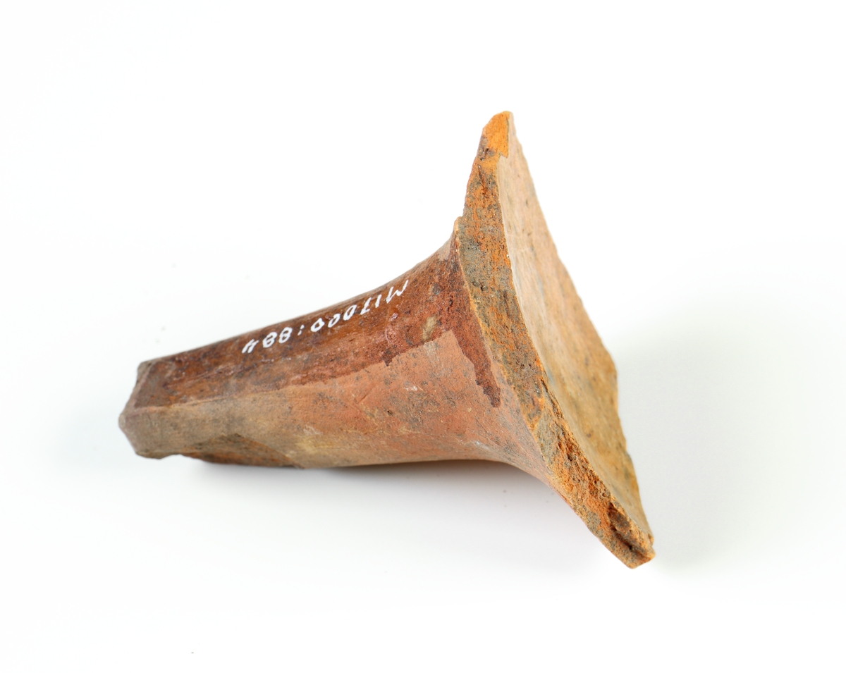 Koniskt ben samt del av kärlbukens insida av yngre rödgods, sannolikt från en trebensgryta. Brun glasyr på grytans insida. Tillverkad i södra Skandinavien.