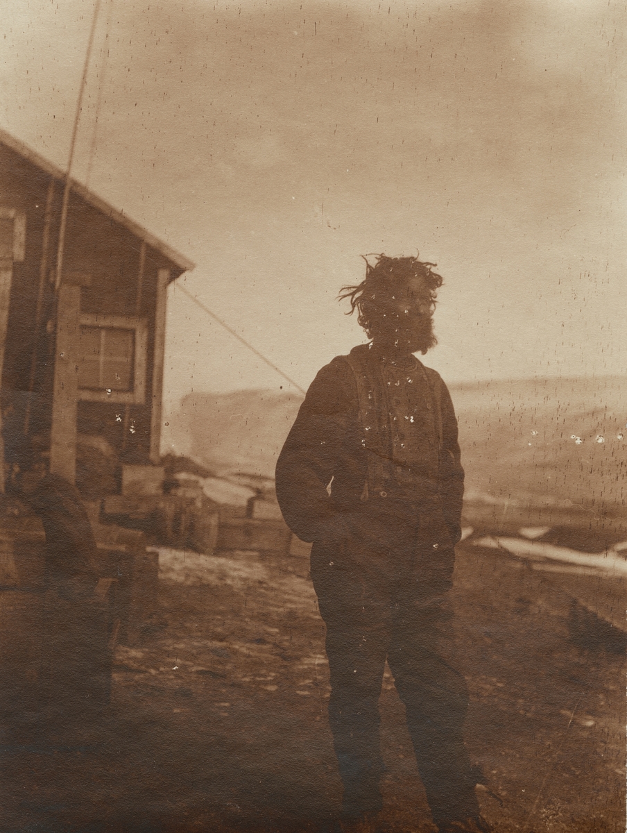 Fotografi från första svenska Antarktisexpeditionen 1901-1904. Motiv av skäggig man stående utanför trähus i Snow Hill.