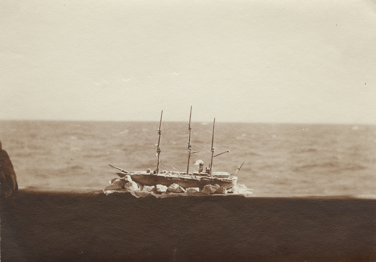 Fotografi från första svenska Antarktisexpeditionen 1901-1904. Motiv av båtmodell som är placerad på en reling vid havet. Modellen liknar fartyget Antarctic.