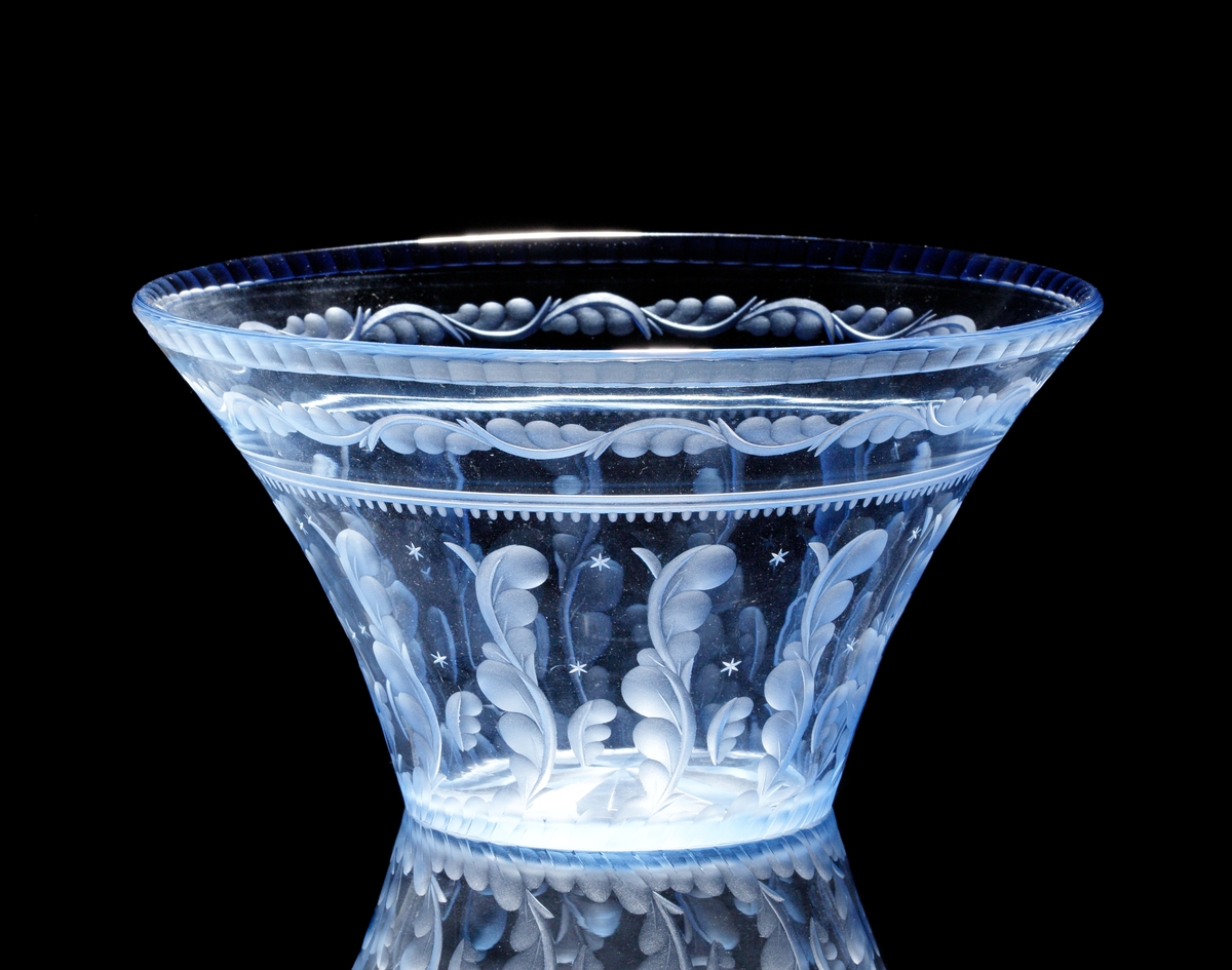 Formgiven av Edward Hald. Ljusblå trattformad skål med fasetterad mynningskant, slipad fjäderformad dekor och
små stjärnor.