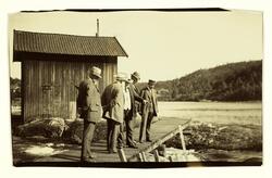 På Skotfoss. Fire menn i dress og med hatt står på en liten 